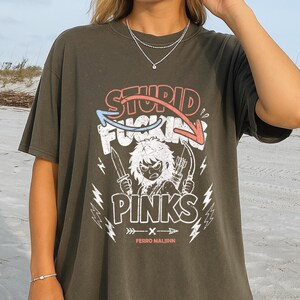 May include: Dark olive green t-shirt with a graphic design. The design features the words "STUPID FUCKING PINKS" in a stylized font, along with an illustration of a character holding knives and arrows. The text "FERRO MALJNN" is below the graphic.