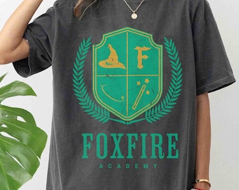 May include: A dark gray t-shirt with a green and yellow crest featuring a wizard's hat, a wand, and a star. The text "Foxfire Academy" is printed below the crest.