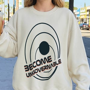 May include: A cream-coloured sweatshirt featuring the text "BECOME UNGOVERNABLE" in red and black. The design includes concentric circles around a black dot. The sweatshirt is a casual, comfortable style.