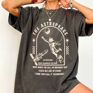 May include: Dark gray t-shirt with a white graphic design featuring an astronaut, a spaceship, and the text "THE ASTROPHAGE". The shirt also includes the text "ENERGY DEVOURER OF THE STARS" and "WHAT WOULD YOU CALL AN ORGANISM THAT EXISTS ON A DIET OF STARS? I THINK YOU'D CALL IT 'ASTROPHAGE.'"