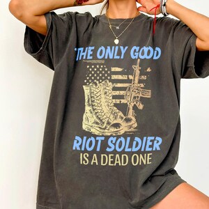 May include: Dark gray oversized t-shirt with a distressed graphic. The graphic features a combat boot, a rifle, and an American flag design, with the text "THE ONLY GOOD RIOT SOLDIER IS A DEAD ONE" in blue.