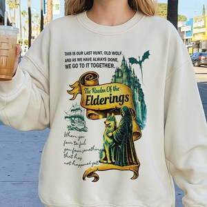 May include: Cream-colored sweatshirt with a graphic design featuring a wolf, a cloaked figure, a castle, and a dragon. The design includes the text "The Realm of the Elderings" and other phrases.
