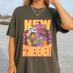 May include: Dark olive green t-shirt with a colorful graphic. The graphic features the words "NEW ACHIEVEMENT" in yellow and pink, along with a cartoon illustration of a man, a cat, and a dragon.