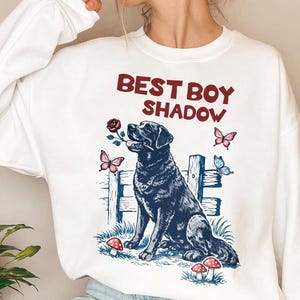 May include: White sweatshirt with a graphic of a black Labrador dog holding a rose in its mouth. The text "BEST BOY SHADOW" is above the dog. The design includes butterflies and mushrooms.