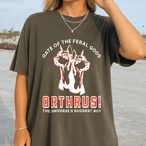 Puede incluir: Camiseta verde oliva oscuro con un gr&aacute;fico de un perro de dos cabezas y el texto "GATE OF THE FERAL GODS" arriba y "ORTHRUS! THE UNIVERSE'S GOODEST BOY" abajo en rojo y blanco. La camiseta es de corte holgado.