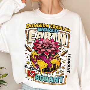 May include: White sweatshirt with a colorful graphic. The graphic features the text "DUNGEON CRAWLER WORLD EARTH" above a globe with a pink flower and the text "Presented by BORANT". Two bullets and small flowers are also included.
