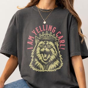 May include: A charcoal gray short-sleeve t-shirt featuring a graphic of a cat wearing a crown and round glasses. The text "I AM YELLING CARL!" is printed in pink above the cat, and the glasses are labeled "PRINCESS DONUT" in small letters.