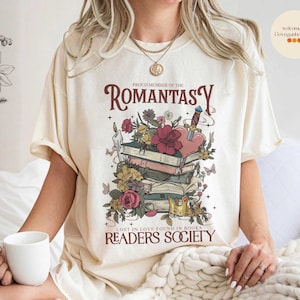May include: A cream-colored t-shirt with a graphic design featuring a stack of books, flowers, and a heart with a sword. The text on the shirt reads "Proud Member of the Romantasy Readers Society. Lost in love found in books."