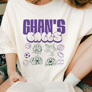 May include: Cream-colored t-shirt with the text "GHAN'S BALLS" in purple bubble letters. Below the text are cartoon illustrations of various sports balls and characters, including a basketball, soccer ball, and volleyball.