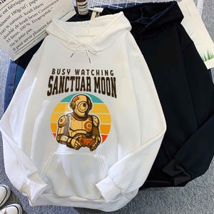 May include: White hoodie with a front pocket and drawstring. The hoodie features a graphic of a robot holding a game controller, set against a sunset design, with the text "BUSY WATCHING SANCTUAR MOON". A black hoodie is next to it.