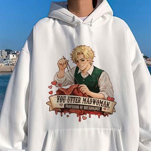 May include: White hoodie with a front pocket and a drawstring hood. The graphic on the front features an illustration of a person with blonde hair wearing a green vest, holding sewing tools, and the text "YOU UTTER MADWOMAN, PROFESSOR OF DREADOLOGY."