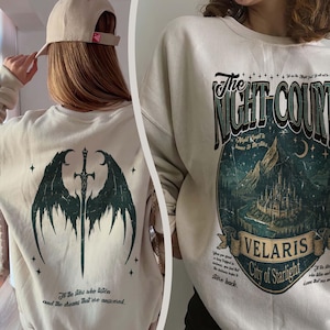 May include: Beige sweatshirt featuring a dark green graphic of a sword and wings on the back, with text that reads "To the stars who listen and the dreams that are answered." The front displays a design with the text "The Night Court" and "Velaris, City of Starlight."