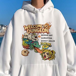 May include: White hooded sweatshirt with a front pocket and a graphic design. The design features the text "WELCOME TO DUNGEON" and cartoon characters. The sweatshirt has white cuffs and a white bottom hem.