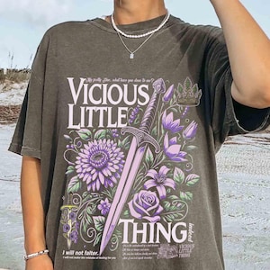 May include: A dark gray t-shirt with a graphic print of a sword, flowers, and the text "Vicious Little Thing" in a vintage style.