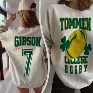 May include: Cream-colored sweatshirt with green lettering and accents. One side displays "GIBSON" and the number "7". The other side features "TOMMEN COLLEGE RUGBY" with a yellow football and shamrocks.