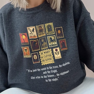 May include: Dark gray sweatshirt featuring a collection of postage stamp-style illustrations in shades of gold, red, and brown. The text "BE WARY BE CLEVER BE GOOD" is printed below the stamps, with a quote below that.