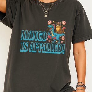 May include: Dark gray t-shirt with the text "MONGO IS APPALLED!" in turquoise. The graphic features a blue dinosaur and a cat wearing a crown, surrounded by donuts. The shirt is a casual, oversized fit.