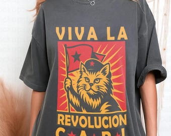 May include: Dark gray t-shirt featuring a cat holding a flag with the text "VIVA LA REVOLUCION CAROL" in yellow. The cat is depicted in a red and yellow graphic, with a star on the flag. The shirt has short sleeves and a relaxed fit.