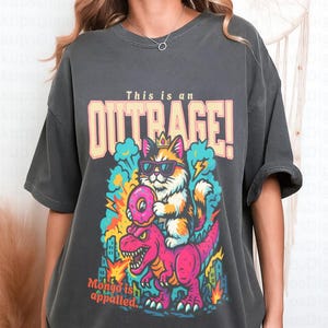 May include: Dark gray t-shirt with a colorful graphic. The graphic features a cat wearing sunglasses and a crown, riding a pink dinosaur. Text on the shirt reads "This is an OUTRAGE!" and "Mongo is appalled."