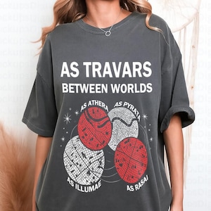 May include: Dark gray t-shirt with the text "AS TRAVARS BETWEEN WORLDS." Below the text are four circular designs in red and white. The shirt is a casual, oversized fit.