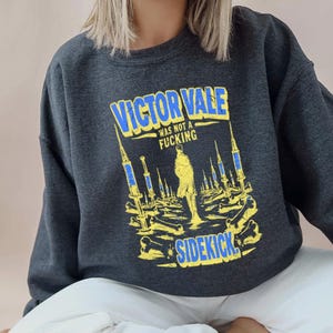 May include: A dark charcoal gray crewneck sweatshirt featuring a graphic print in yellow and blue. The text reads VICTOR VALE WAS NOT A FUCKING SIDEKICK, surrounded by illustrations of syringes and bones arranged in a landscape pattern.