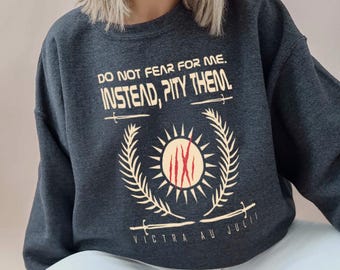 Pode incluir: Camisola cinza escuro com o texto "DO NOT FEAR FOR ME. INSTEAD, PITY THEM." e um desenho de sol com um centro vermelho. O desenho é emoldurado por ramos de louro e espadas cruzadas. O texto "VICTRA AU JULI" está em baixo.
