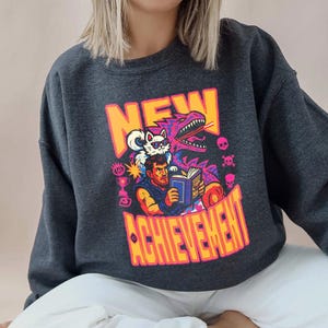 May include: Dark gray sweatshirt featuring a colorful graphic with the words "NEW ACHIEVEMENT". The graphic includes a man reading a book, a cat, a dinosaur, and other gaming-related icons. The sweatshirt is a casual, comfortable style.
