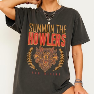 May include: Dark gray t-shirt with the text "SUMMON THE HOWLERS" in orange and red. Below the text is a graphic of a three-headed wolf with a laurel wreath and the words "RED RISING".