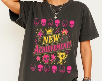 May include: Dark gray t-shirt with a graphic design featuring pink skulls, a gold crown, lightning bolts, and the text "NEW ACHIEVEMENT!" in yellow and pink. A trophy and star are also included in the design.