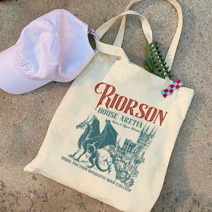 May include: A white canvas tote bag featuring a green dragon and a castle graphic. The text on the bag reads "Riorson House Aretia Riders & Tygers Welcome Rebel Faction Basgiath War College".