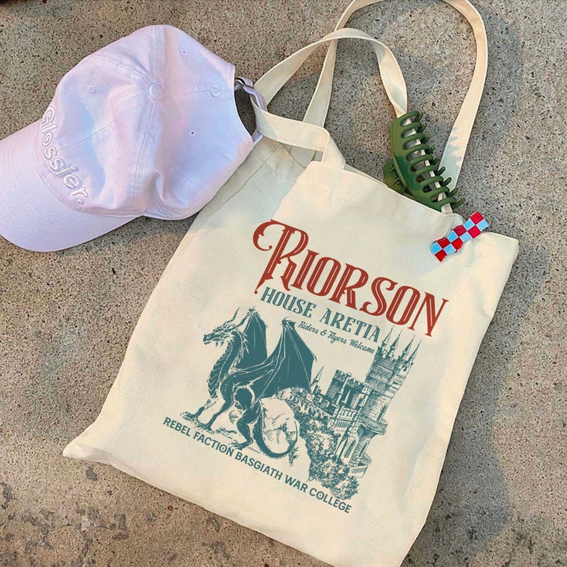 May include: A white canvas tote bag featuring a green dragon and a castle graphic. The text on the bag reads "Riorson House Aretia Riders & Tygers Welcome Rebel Faction Basgiath War College".