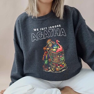 May include: Dark gray sweatshirt with the text "WE JUST IGNORE AGATHA" in white. The graphic depicts a person pushing a shopping cart filled with groceries and a pink flamingo. The cart has the IKEA logo.