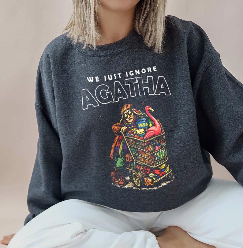 May include: Dark gray sweatshirt with the text "WE JUST IGNORE AGATHA" in white. The graphic depicts a person pushing a shopping cart filled with groceries and a pink flamingo. The cart has the IKEA logo.