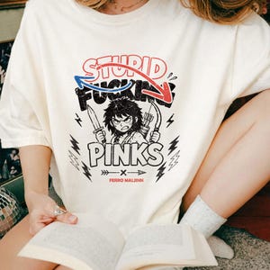 May include: Cream-colored t-shirt with a graphic featuring the text "STUPID FUCKING PINKS" above an illustration of an angry character holding a knife and bow and arrows. The shirt also has the text "FERRO MALJINN".