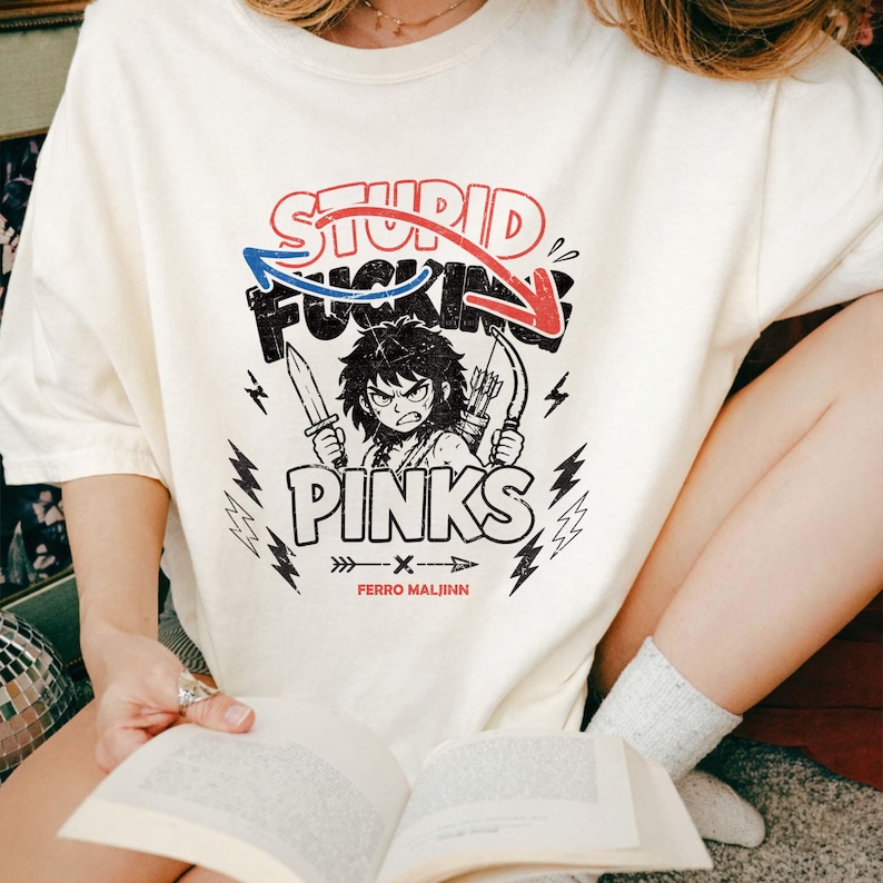 May include: Cream-colored t-shirt with a graphic featuring the text "STUPID FUCKING PINKS" above an illustration of an angry character holding a knife and bow and arrows. The shirt also has the text "FERRO MALJINN".