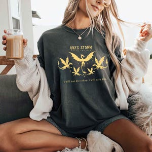 May include: A black long-sleeved shirt with a yellow graphic of a dragon and the text "ONYX STORM" and "Brave the Dark". The text "I will not die today. I will save him." is printed below the graphic.