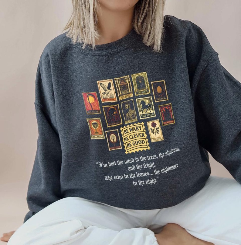 May include: Dark gray sweatshirt featuring a collection of postage stamp-style illustrations in shades of gold, red, and brown. The text "BE WARY BE CLEVER BE GOOD" is printed below the stamps, with a quote below that.