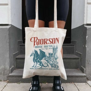May include: A white canvas tote bag with a graphic design featuring a dragon and a castle. The text on the bag reads "Riorson House Aretia Riders & Flyers Welcome Rebel Faction Basgiath War College".