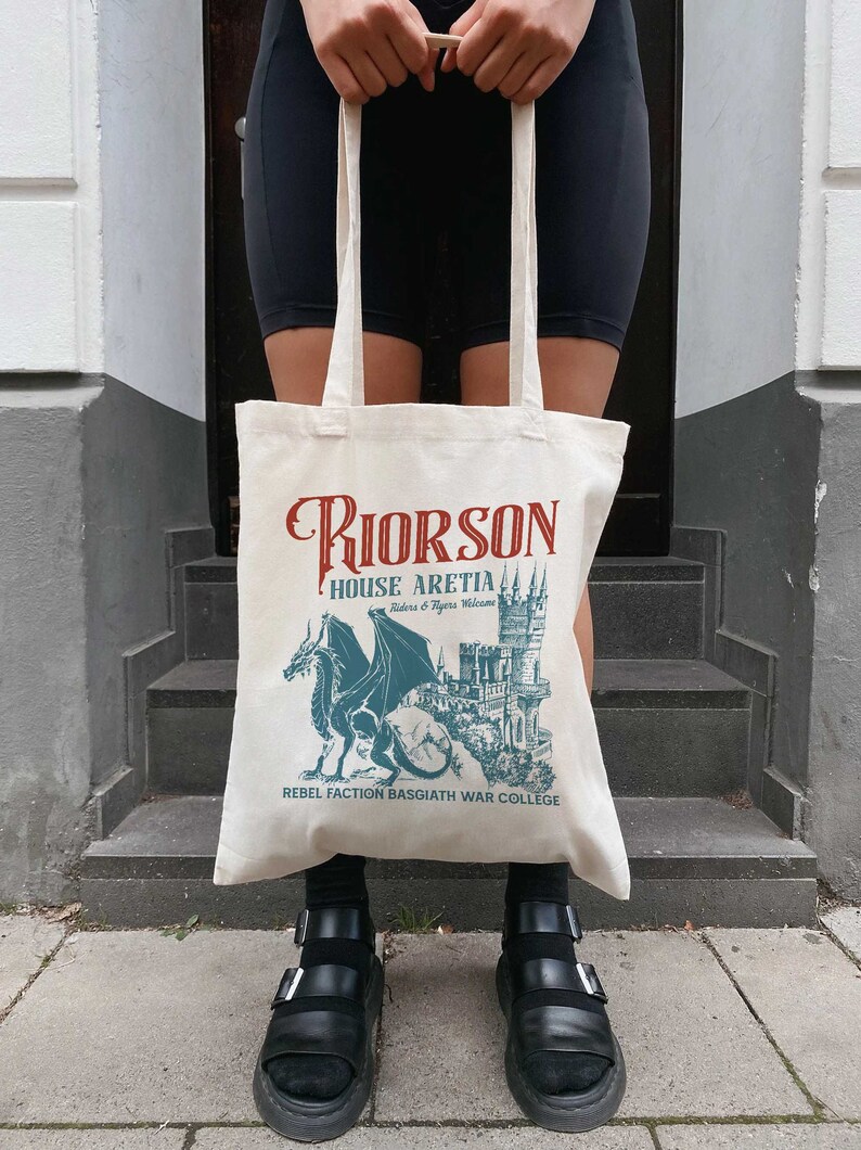 May include: A white canvas tote bag with a graphic design featuring a dragon and a castle. The text on the bag reads "Riorson House Aretia Riders & Flyers Welcome Rebel Faction Basgiath War College".