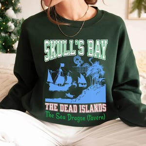 May include: Dark green sweatshirt with a graphic featuring the text "SKULL'S BAY" above a blue illustration of sailing ships and an island. Below the image, the text reads "THE DEAD ISLANDS" and "The Sea Dragon (Tavern)".