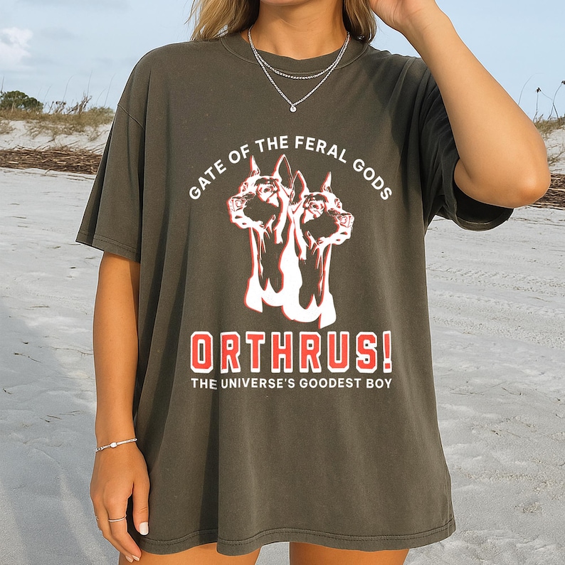 Puede incluir: Camiseta verde oliva oscuro con un gr&aacute;fico de un perro de dos cabezas y el texto "GATE OF THE FERAL GODS" arriba y "ORTHRUS! THE UNIVERSE'S GOODEST BOY" abajo en rojo y blanco. La camiseta es de corte holgado.