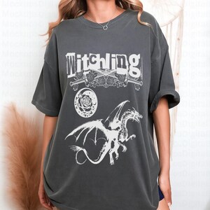 May include: Dark grey t-shirt featuring the word "Witchling" above a design of crossed swords, roses, a crescent moon, and a dragon. The graphic is in white. The shirt has short sleeves and a relaxed fit.