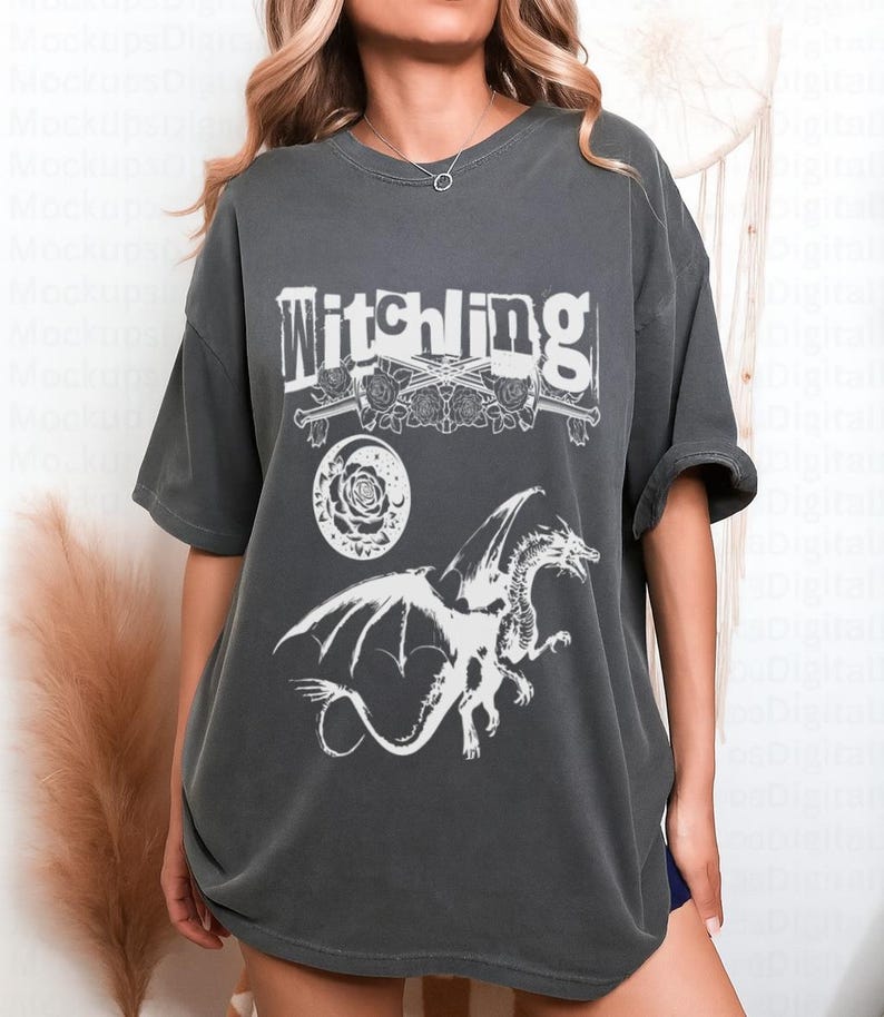 May include: Dark grey t-shirt featuring the word "Witchling" above a design of crossed swords, roses, a crescent moon, and a dragon. The graphic is in white. The shirt has short sleeves and a relaxed fit.