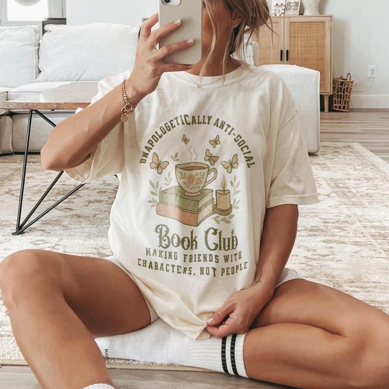 May include: Cream-colored t-shirt with the text "Unapologetically Anti-Social Book Club" and a graphic of a teacup, books, butterflies, and a candle. The shirt also has the phrase "Making Friends With Characters, Not People."