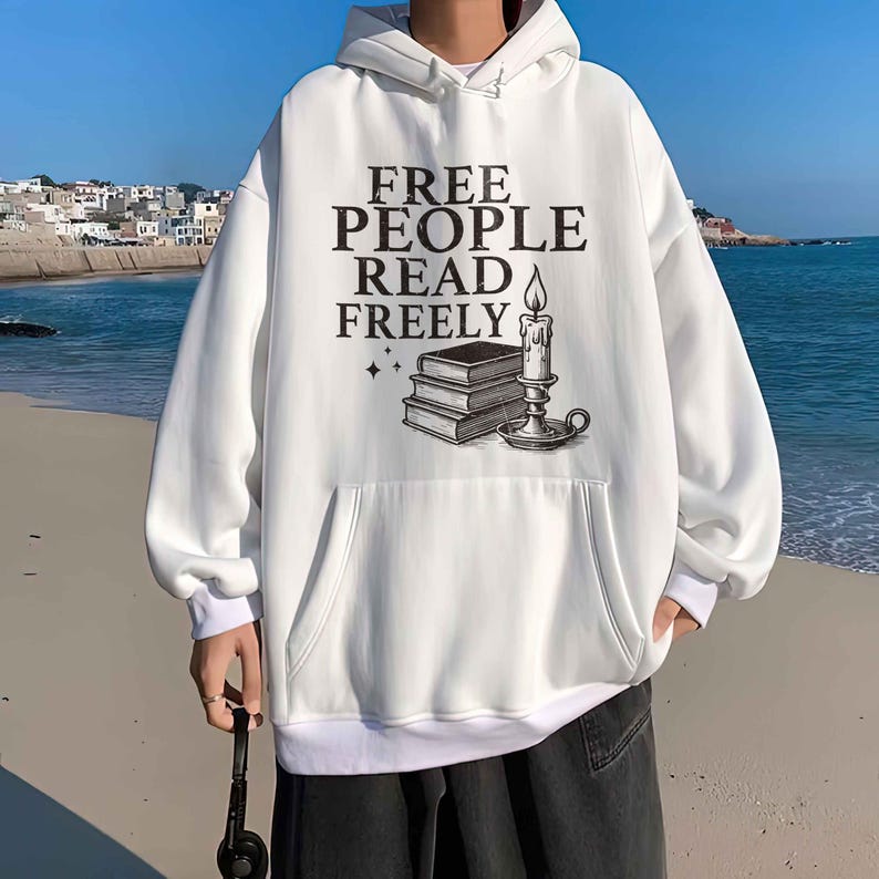 Op de afbeelding: Witte hoodie met een voorzak en de tekst "FREE PEOPLE READ FREELY" in zwart. De afbeelding bevat een stapel boeken, een kaars en sterren. De hoodie heeft een capuchon met trekkoord en geribde manchetten.