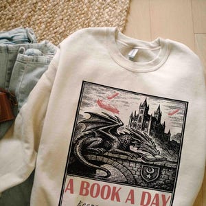 May include: Cream-colored sweatshirt with a graphic of a dragon, castle, and books. The graphic is in black and white with red accents. Text on the sweatshirt reads "A BOOK A DAY keeps..."