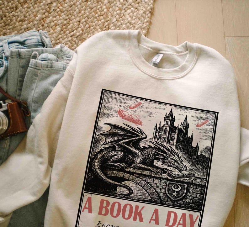 May include: Cream-colored sweatshirt with a graphic of a dragon, castle, and books. The graphic is in black and white with red accents. Text on the sweatshirt reads "A BOOK A DAY keeps..."
