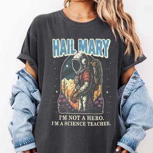 May include: Dark gray t-shirt with the text "HAIL MARY" in blue and white. The shirt features a graphic of an astronaut in a spacesuit, with the text "I'M NOT A HERO. I'M A SCIENCE TEACHER."