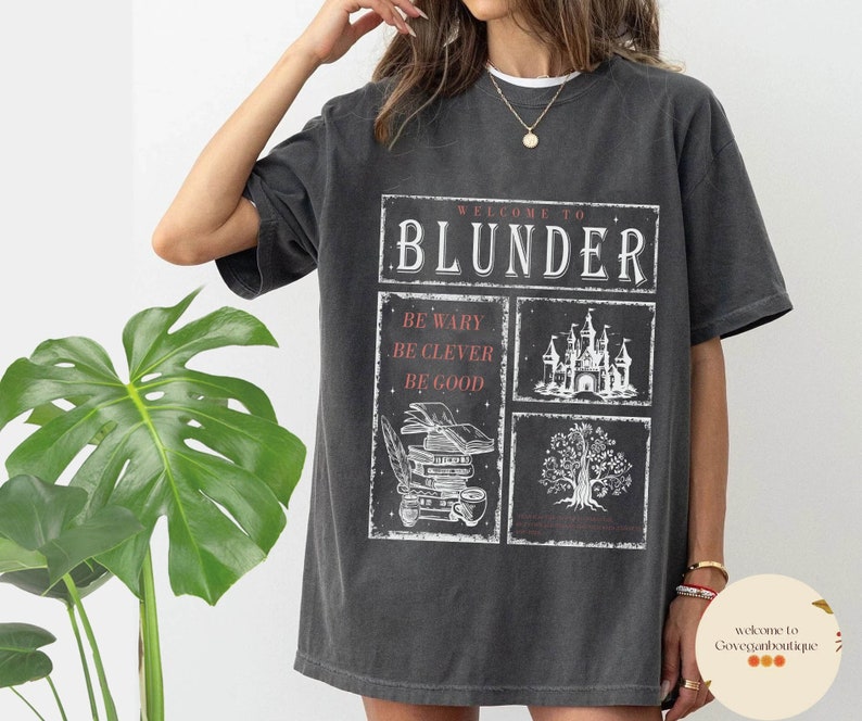 Op de afbeelding: Een oversized donkergrijs T-shirt met een grafische print die "Welkom bij Blunder" zegt met een kasteel, een boom en boeken. De tekst onder de grafische print zegt "Wees voorzichtig, wees slim, wees goed."