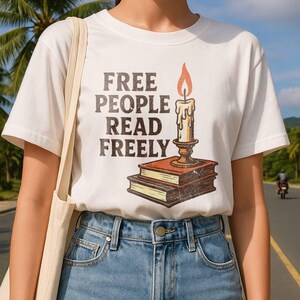 Może przedstawiać: Biała koszulka z napisem "FREE PEOPLE READ FREELY" w brązowych literach. Koszulka przedstawia grafikę świecy na dw&oacute;ch książkach. Świeca ma ż&oacute;łty płomień i jest w złotym uchwycie.